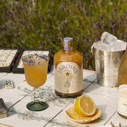 Flasche Jardins alkoholfreies Getränk mit gelbem Inhalt, Cocktailglas mit Eis und Blüten, Zitrone auf weißem Kacheltisch.
