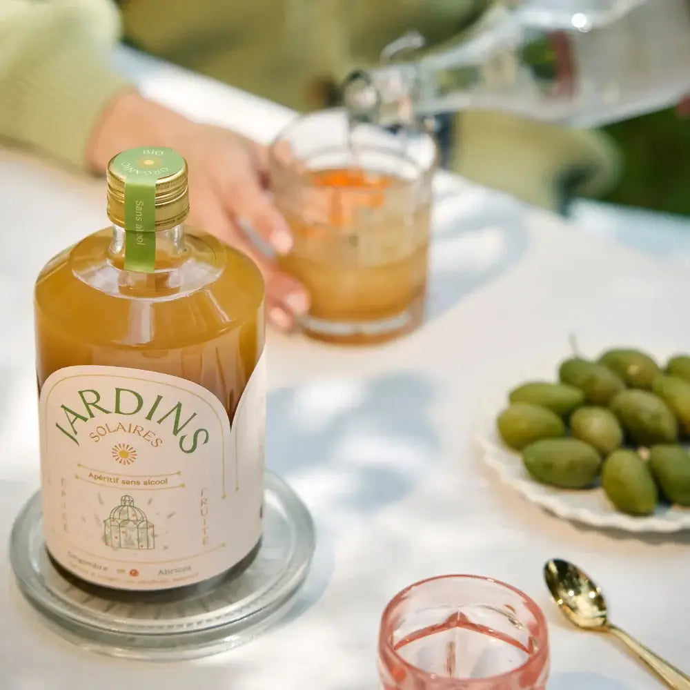 Flasche Jardins alkoholfreies Getränk 70 cl mit grünem Korken, Glas mit Eis und Olivenschale auf weißem Tisch.