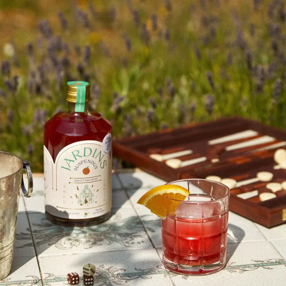 70 cl Flasche alkoholfreier roter Jardins-Cocktail mit Rosmarinzweig, Glas mit Orangenscheibe und Spielbrett im Hintergrund.
