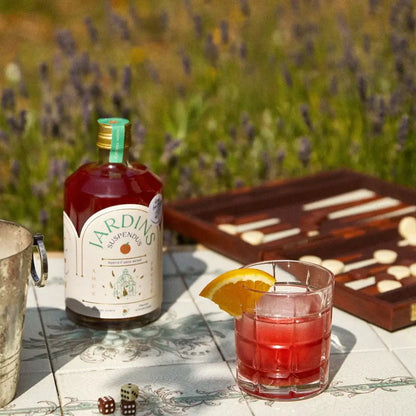 70 cl Flasche alkoholfreier roter Jardins-Cocktail mit Rosmarinzweig, Glas mit Orangenscheibe und Spielbrett im Hintergrund.