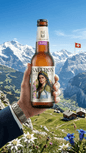 Hand hält Flasche Cyrus Saffron Alkoholfreies Safran Craft Beer vor Bergkulisse mit Schweizer Flagge.