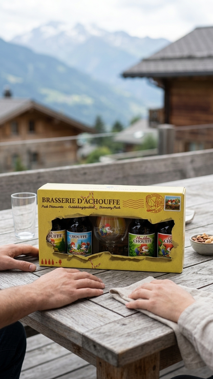 Geschenkbox mit drei Flaschen La Chouffe Bier auf rustikalem Holztisch vor Alpenhintergrund.